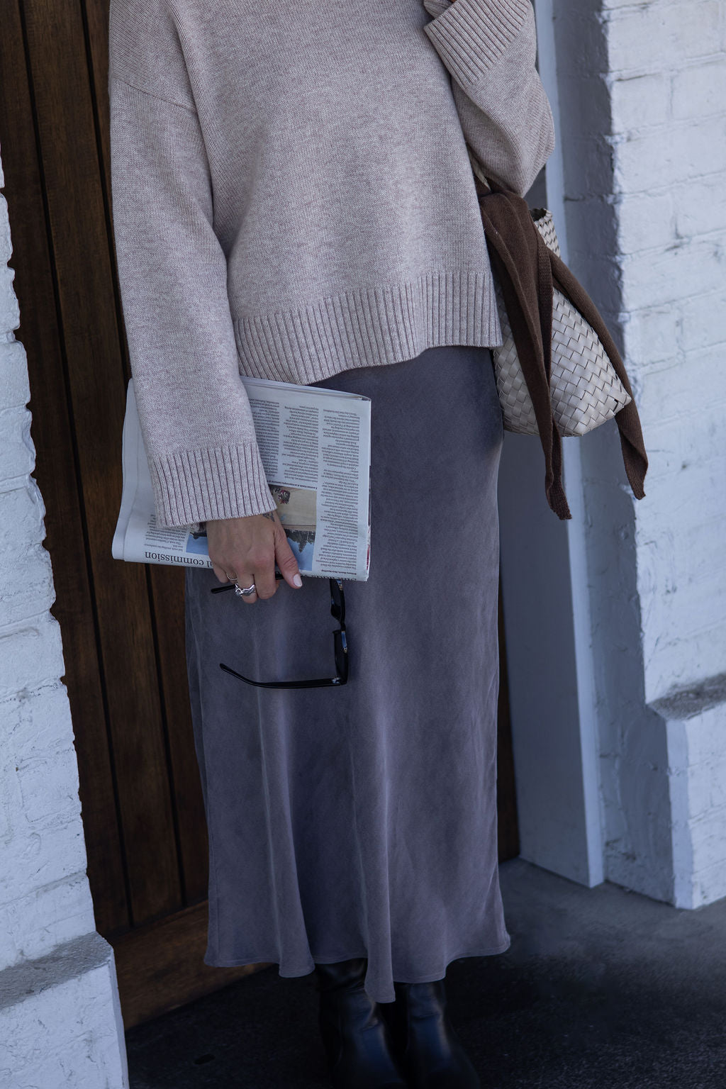 Person wearing a beige sweater and gray skirt holding a newspaper, standing against a white brick wall.