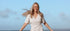 Woman in a white dress standing on a beach with a clear blue sky.