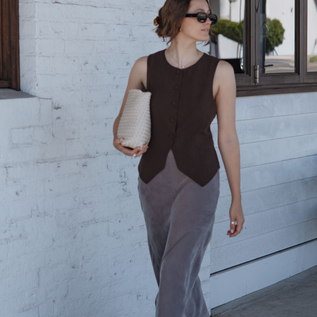 Woman in a sleeveless brown top and gray pants standing against a white wall.