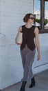 Woman in a sleeveless brown top and gray pants standing against a white wall.