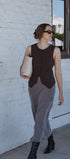 Woman in a sleeveless brown top and gray pants standing against a white wall.