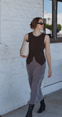 Woman in a sleeveless brown top and gray pants standing against a white wall.