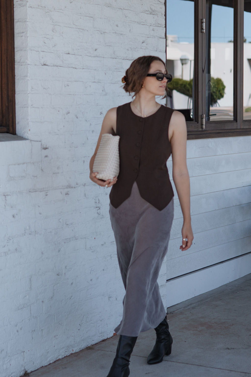 Woman in a sleeveless brown top and gray pants standing against a white wall.