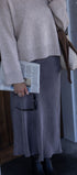 Person wearing a beige sweater and gray skirt holding a newspaper, standing against a white brick wall.