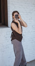 Person taking a mirror selfie wearing a brown top and gray pants against a white brick wall.