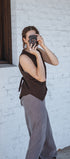 Person taking a mirror selfie wearing a brown top and gray pants against a white brick wall.