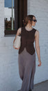 Woman wearing a brown vest over a long dress, holding a white bag, against a white brick wall.