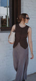 Woman wearing a brown vest over a long dress, holding a white bag, against a white brick wall.