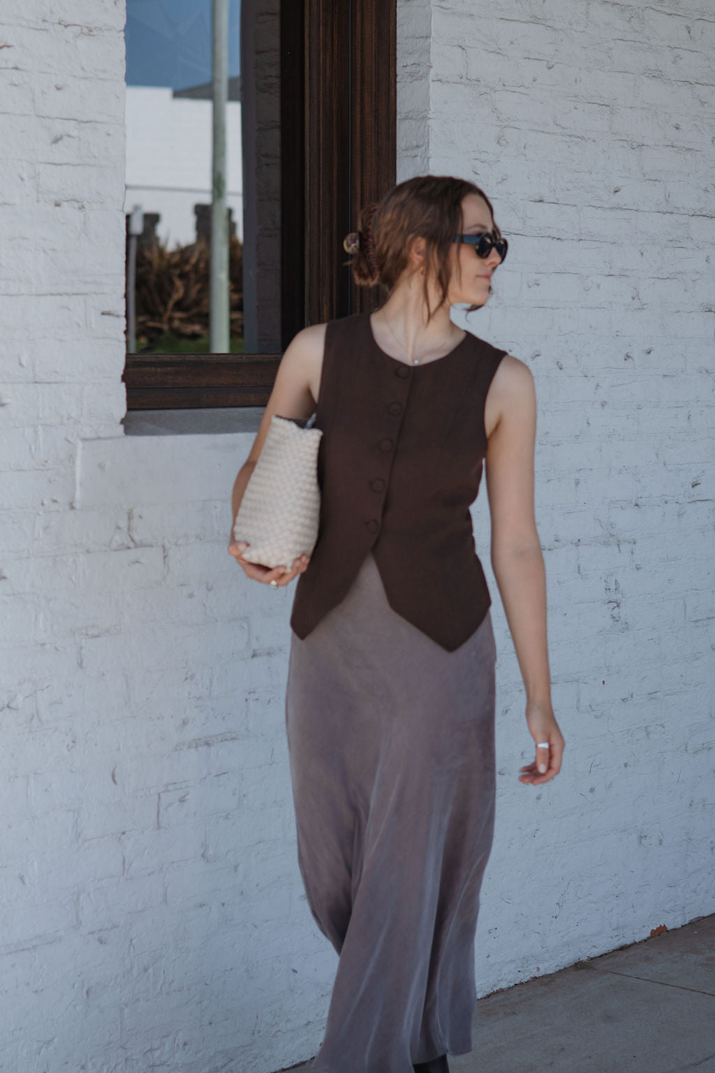 Woman wearing a brown vest over a long dress, holding a white bag, against a white brick wall.