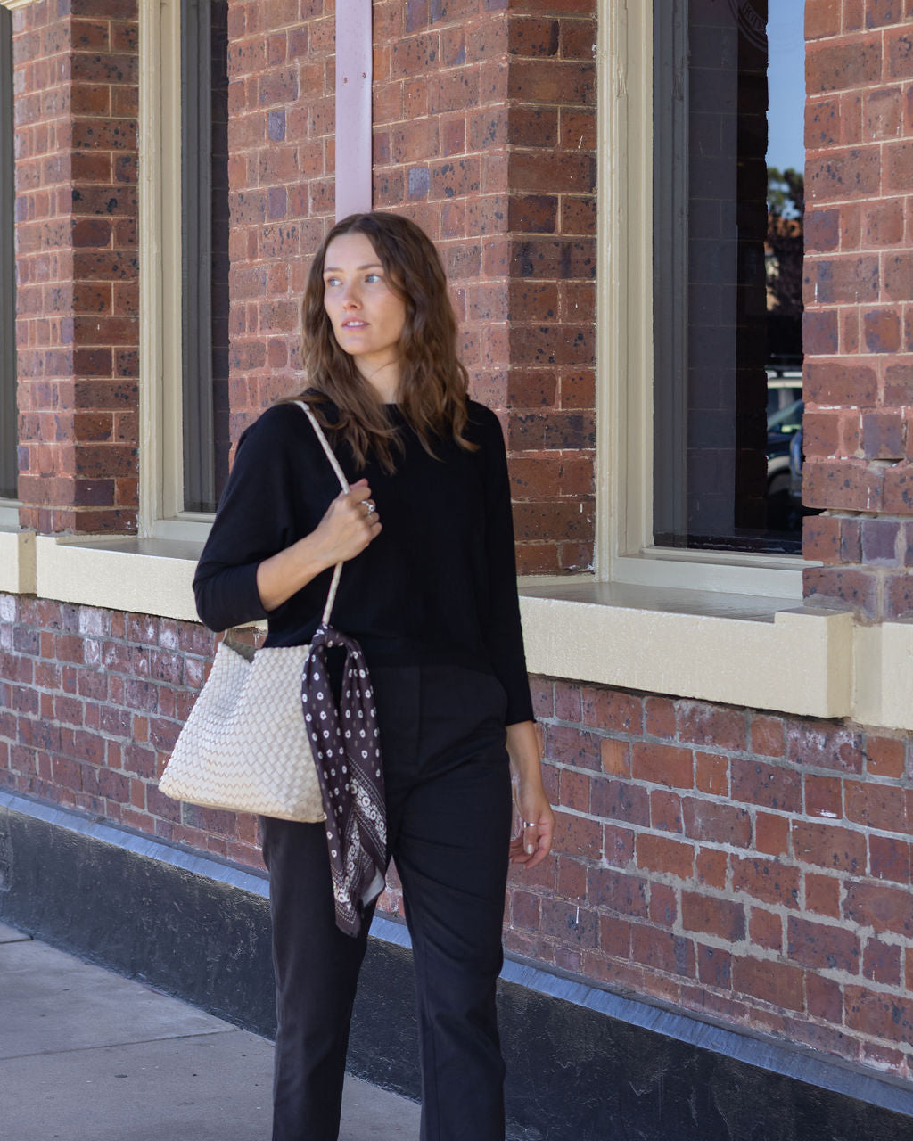 Woman holding a patterned scarf and a white handbag against a brick building background