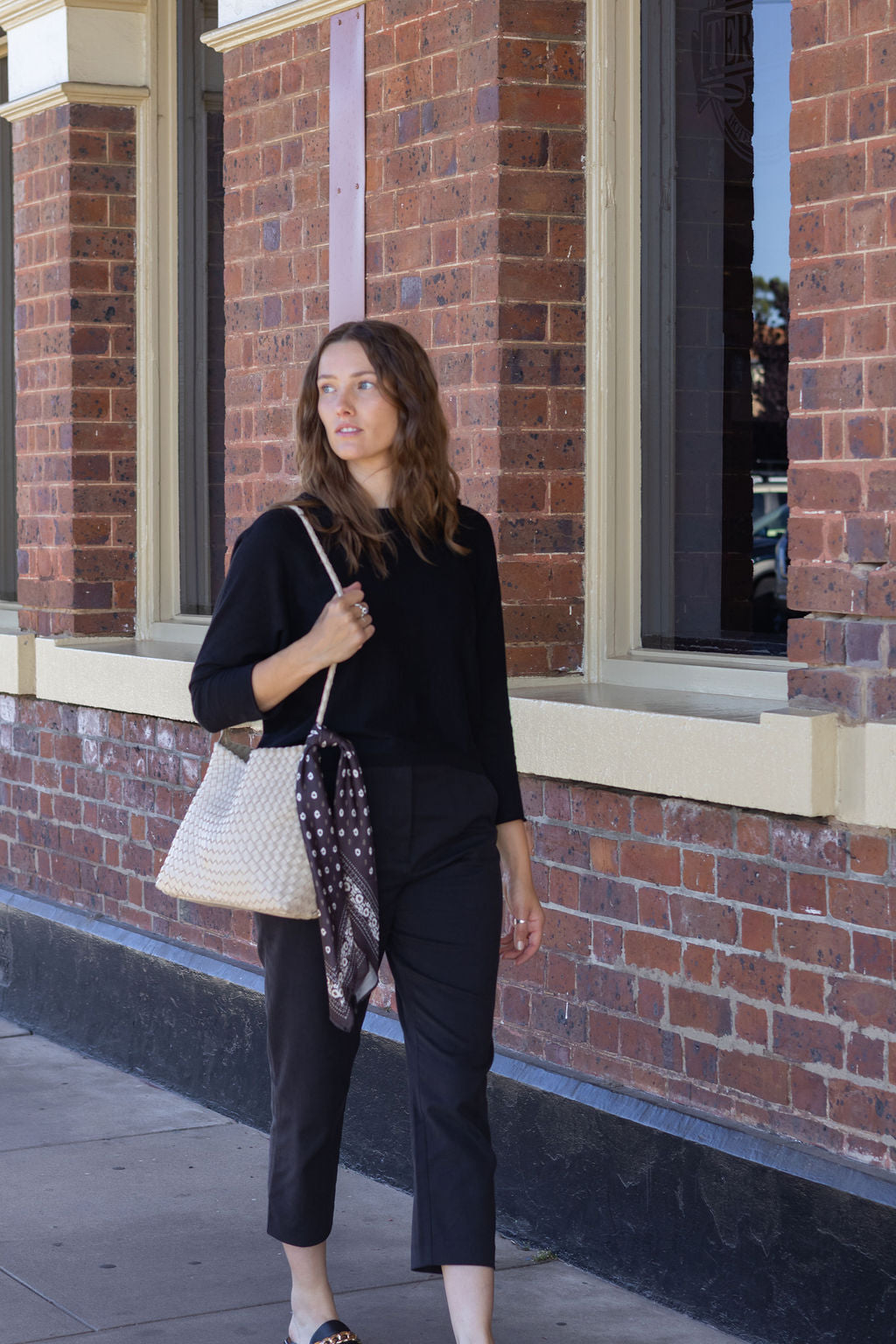 Woman holding a patterned scarf and a white handbag against a brick building background
