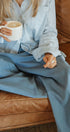 Person sitting on a brown leather couch holding a white mug, wearing a light blue shirt and pants.