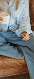 Person sitting on a brown leather couch holding a white mug, wearing a light blue shirt and pants.