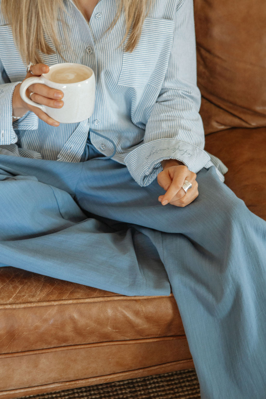 Person sitting on a brown leather couch holding a white mug, wearing a light blue shirt and pants.