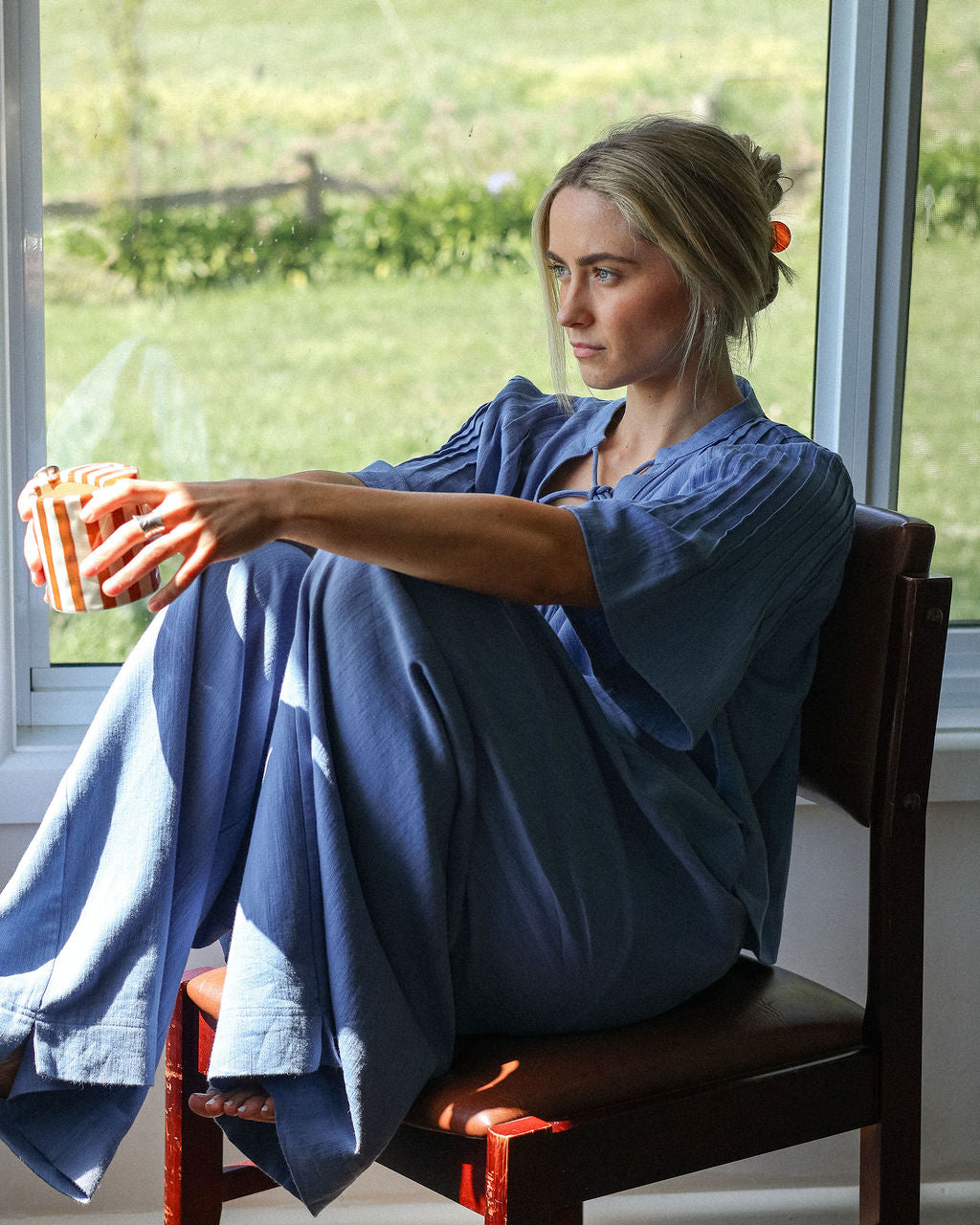 Woman sitting on a chair by a window holding a bowl, with a scenic view outside.