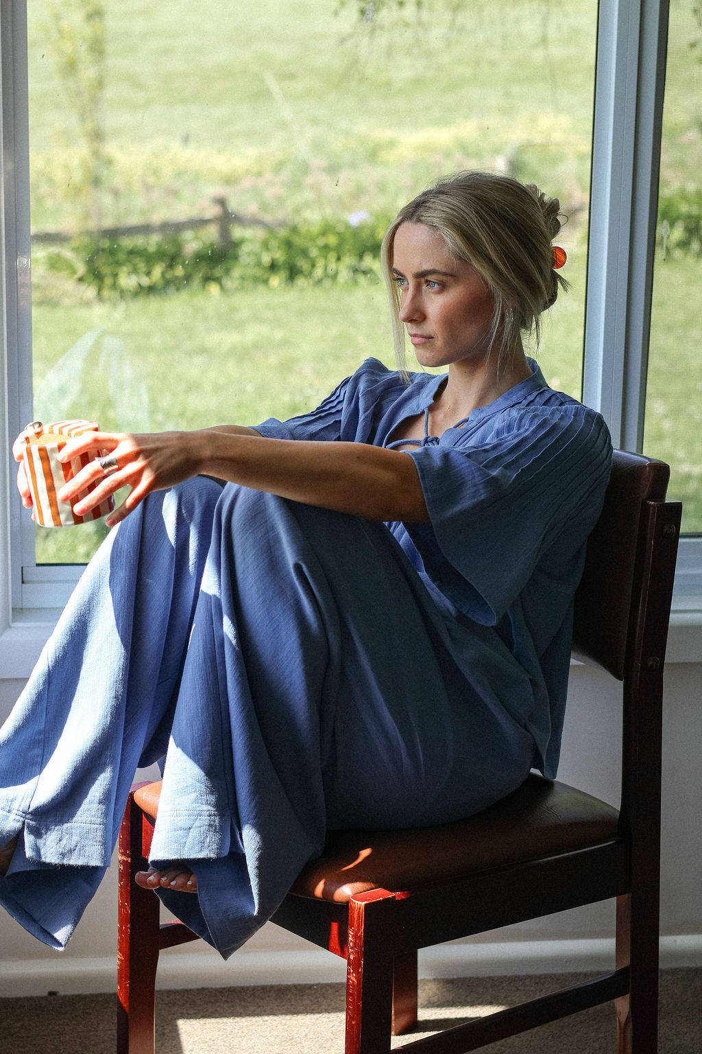 Woman sitting on a chair by a window holding a bowl, with a scenic view outside.