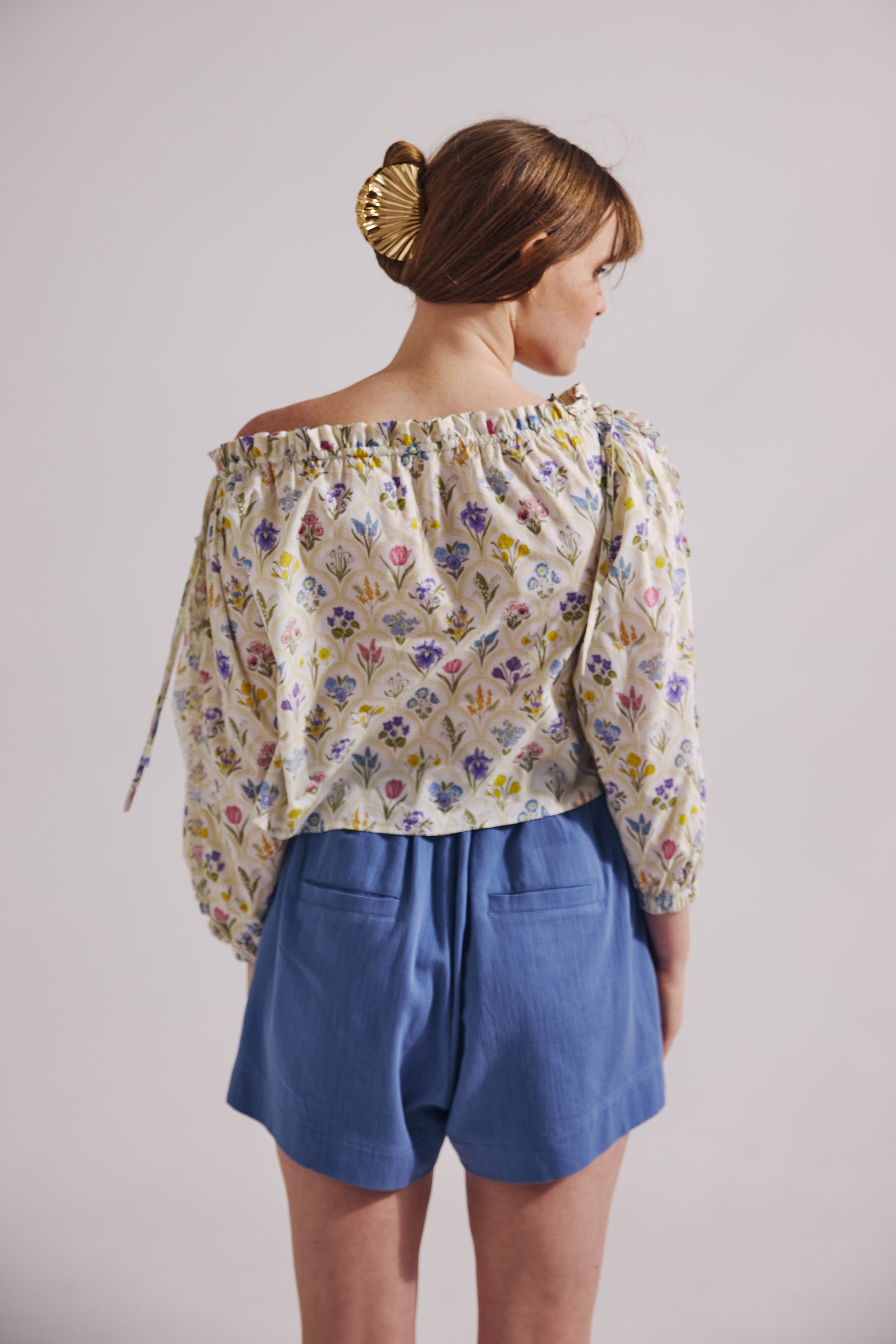 Woman wearing a floral off-shoulder top and blue shorts against a plain background