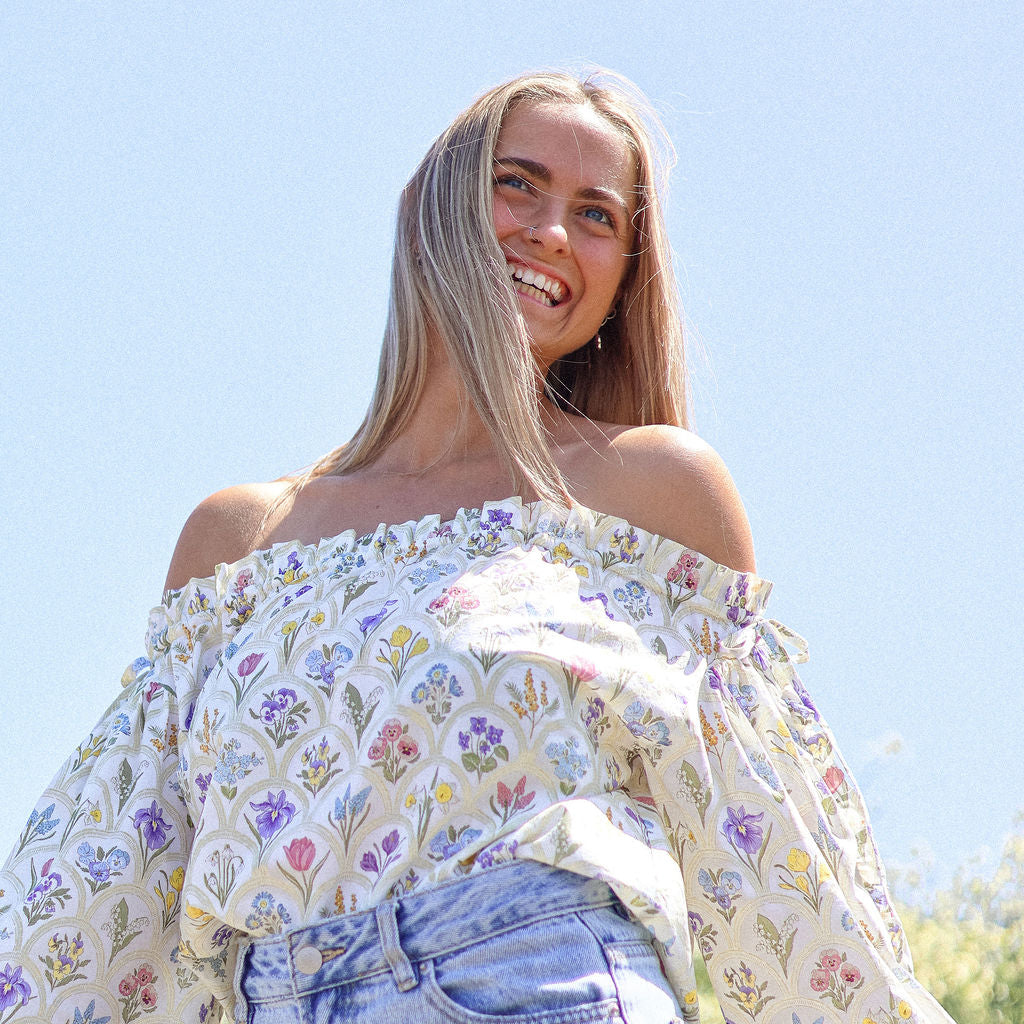 Woman wearing a floral off-shoulder top and jeans against a clear blue sky.