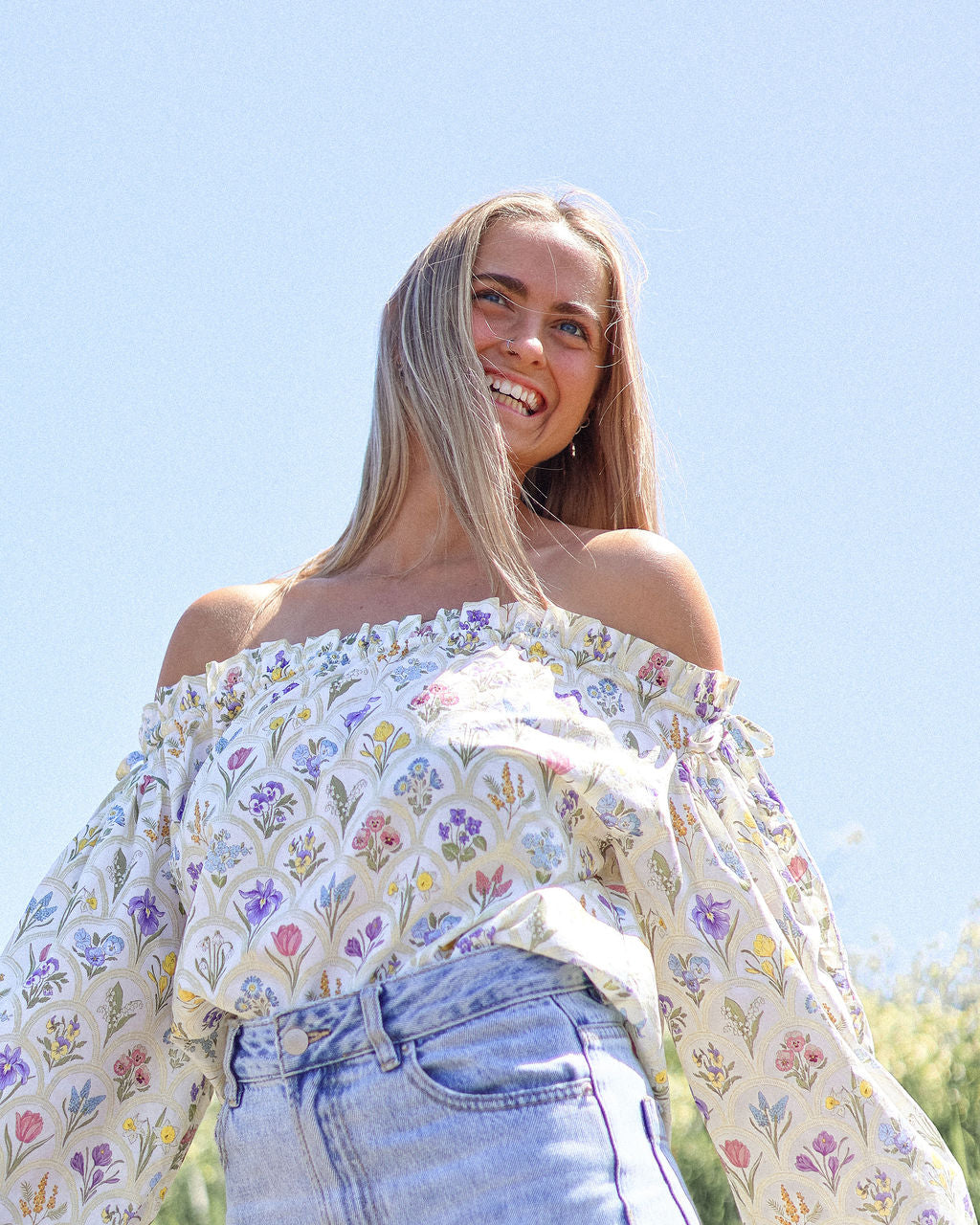 Woman wearing a floral off-shoulder top and jeans against a clear blue sky.
