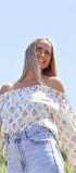 Woman wearing a floral off-shoulder top and jeans against a clear blue sky.