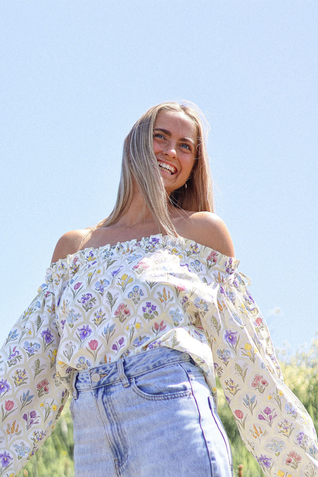 Woman wearing a floral off-shoulder top and jeans against a clear blue sky.