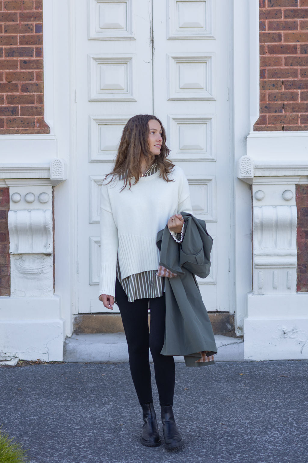 Woman standing outside a building with large white doors, wearing a white sweater and black pants.