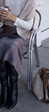 Person sitting at a table with a cup of coffee, wearing a beige sweater and black boots, with a handbag on the ground.