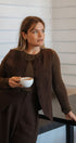 Woman in a brown outfit holding a white cup, standing indoors.