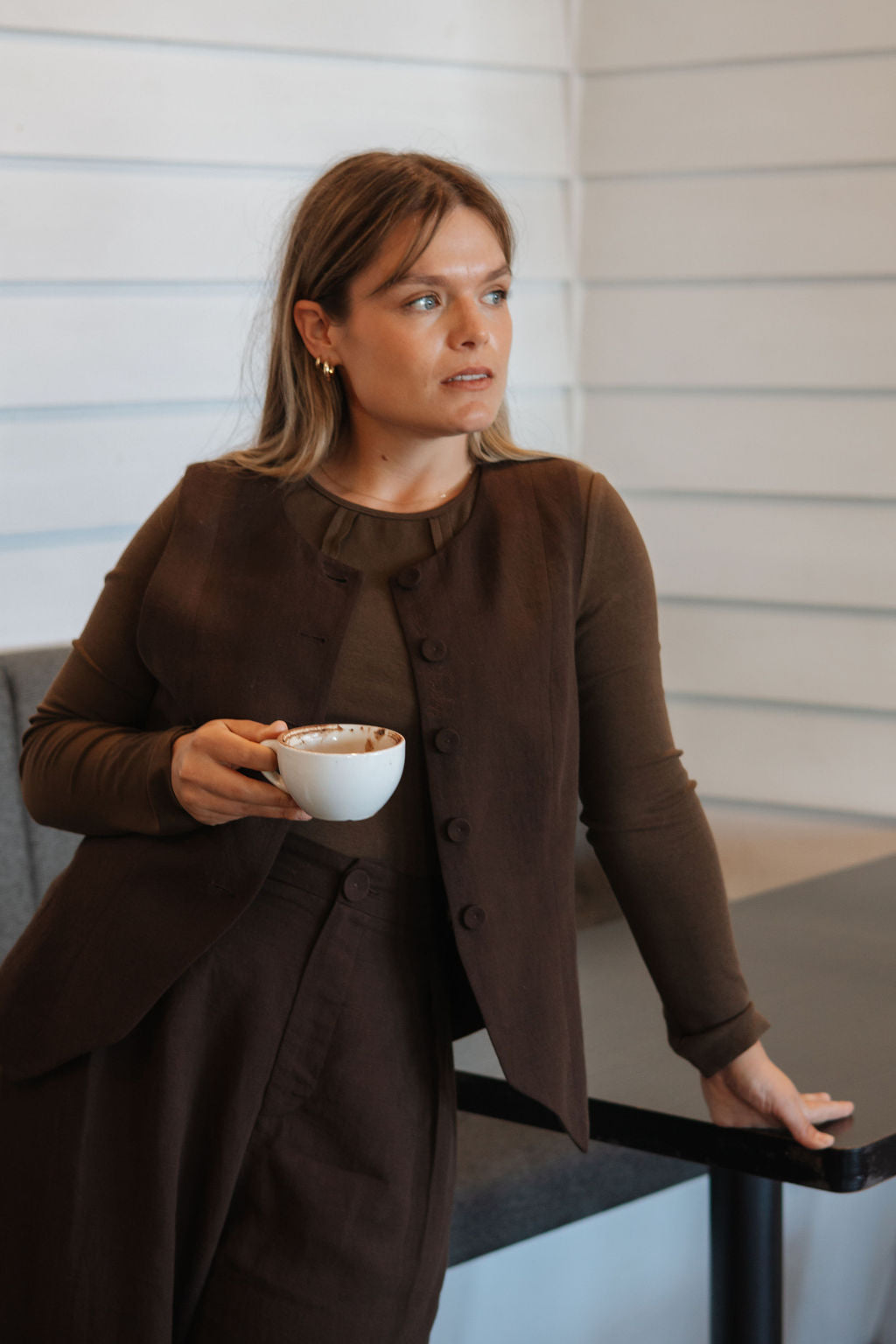 Woman in a brown outfit holding a white cup, standing indoors.