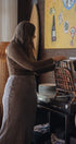 Woman in a kitchen reaching for a plate on a shelf with a colorful painting in the background.
