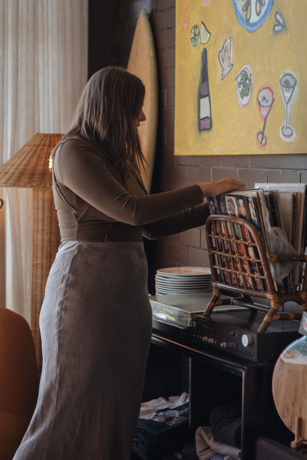Woman in a kitchen reaching for a plate on a shelf with a colorful painting in the background.