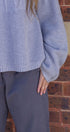 Person wearing a textured blue sweater and dark pants against a brick wall.