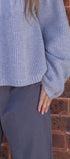 Person wearing a textured blue sweater and dark pants against a brick wall.