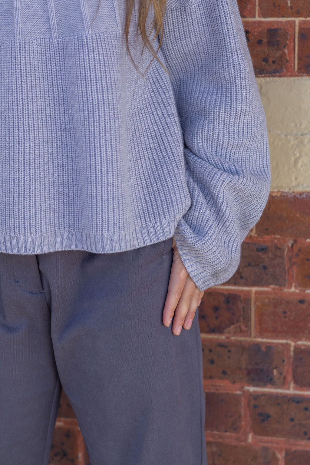 Person wearing a textured blue sweater and dark pants against a brick wall.