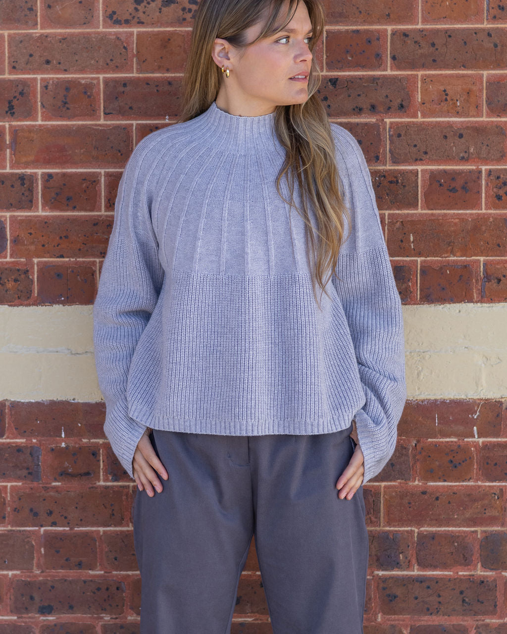 Woman wearing a light blue ribbed sweater and gray pants against a brick wall.