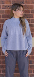 Woman wearing a light blue ribbed sweater and gray pants against a brick wall.