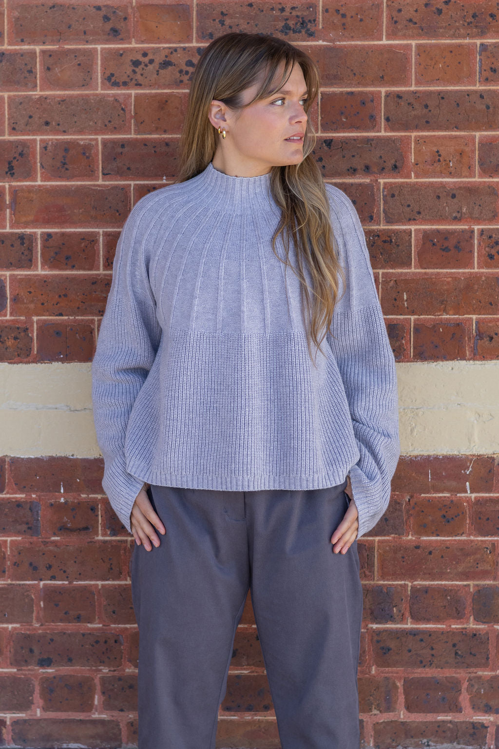 Woman wearing a light blue ribbed sweater and gray pants against a brick wall.