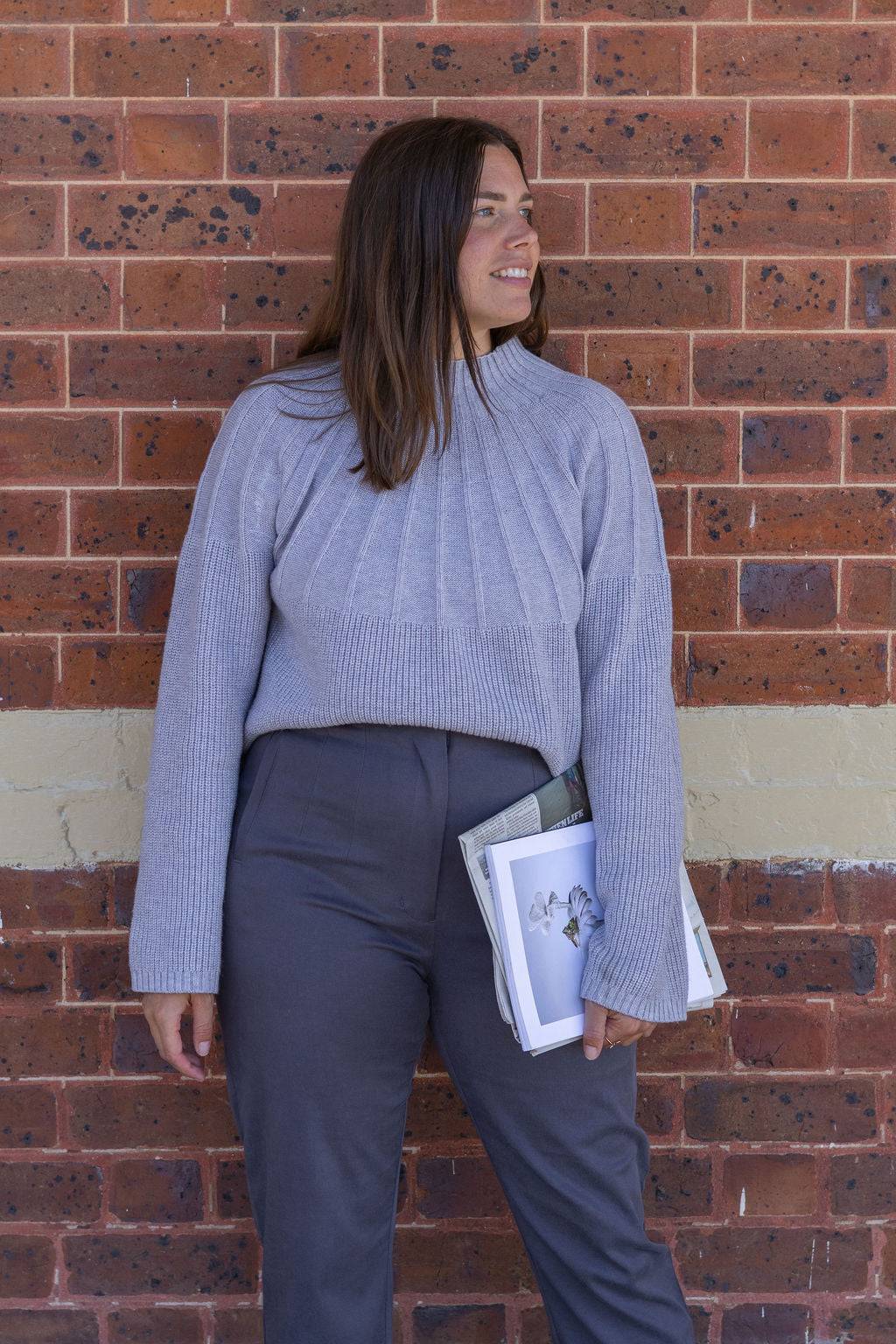 Woman in a gray sweater and dark pants standing against a brick wall