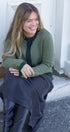 Woman sitting on a step wearing a green sweater and dark pants, smiling.