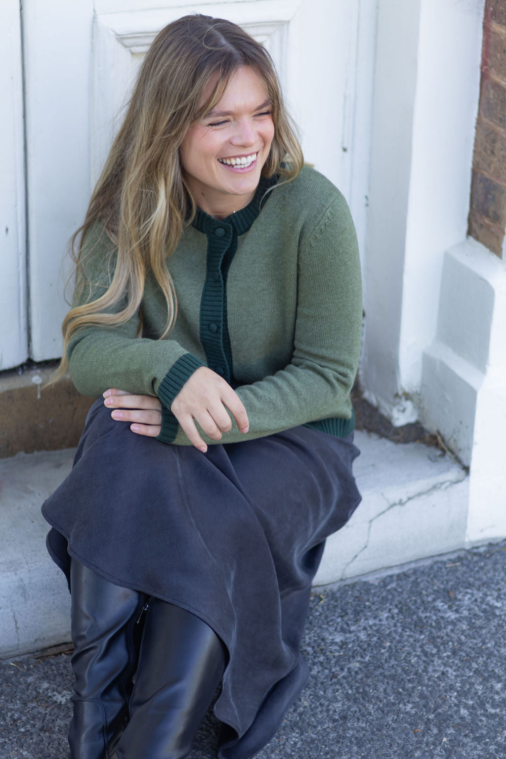 Woman sitting on a step wearing a green sweater and dark pants, smiling.
