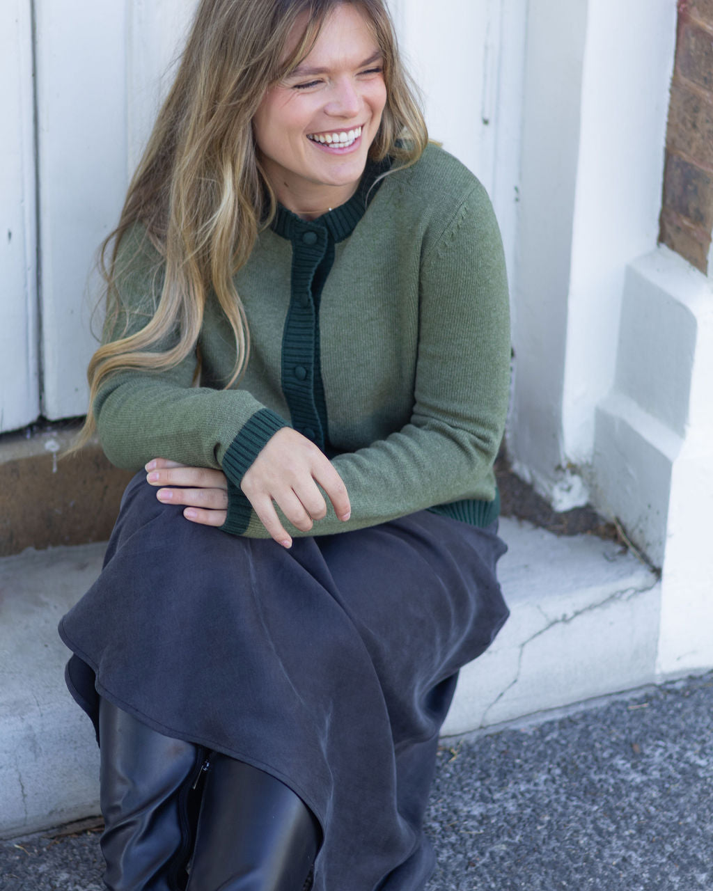 Woman sitting on a step wearing a green sweater and dark pants, smiling.