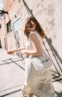 Woman in a white dress holding a white bag with text, standing against a textured wall.