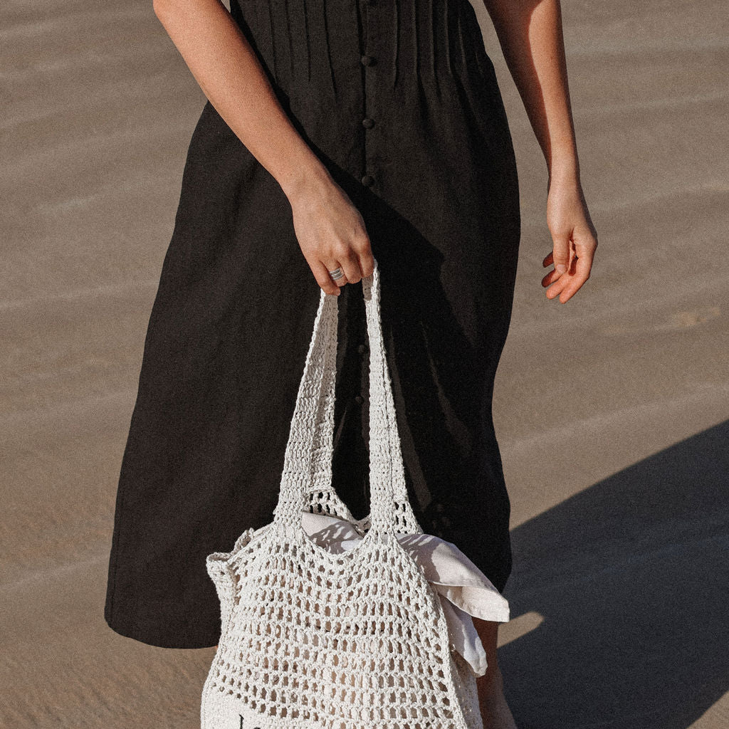 Person holding a white woven bag with 'LOVE OF IVY' text on a sandy surface