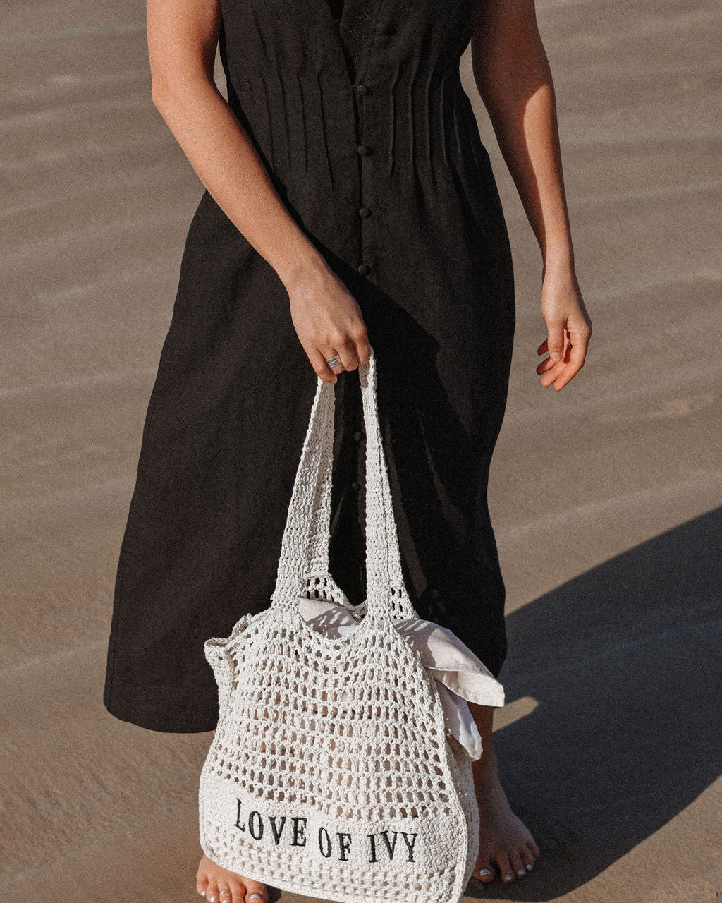 Person holding a white woven bag with 'LOVE OF IVY' text on a sandy surface