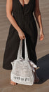 Person holding a white woven bag with 'LOVE OF IVY' text on a sandy surface