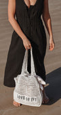 Person holding a white woven bag with 'LOVE OF IVY' text on a sandy surface