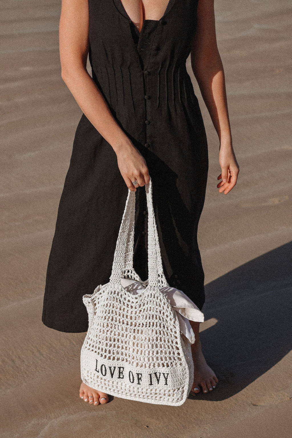 Person holding a white woven bag with 'LOVE OF IVY' text on a sandy surface