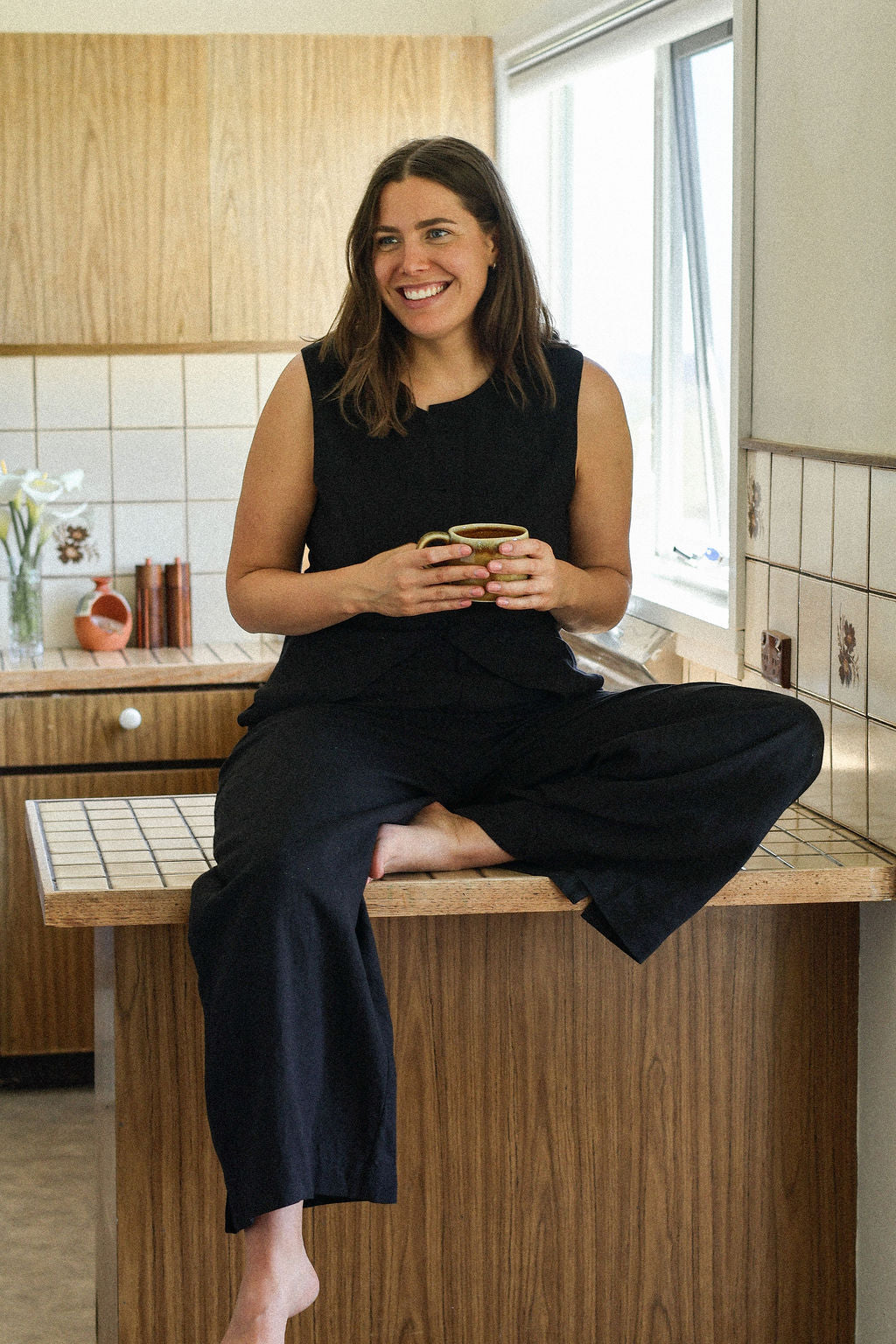 Woman sitting on a kitchen counter holding a cup, smiling.