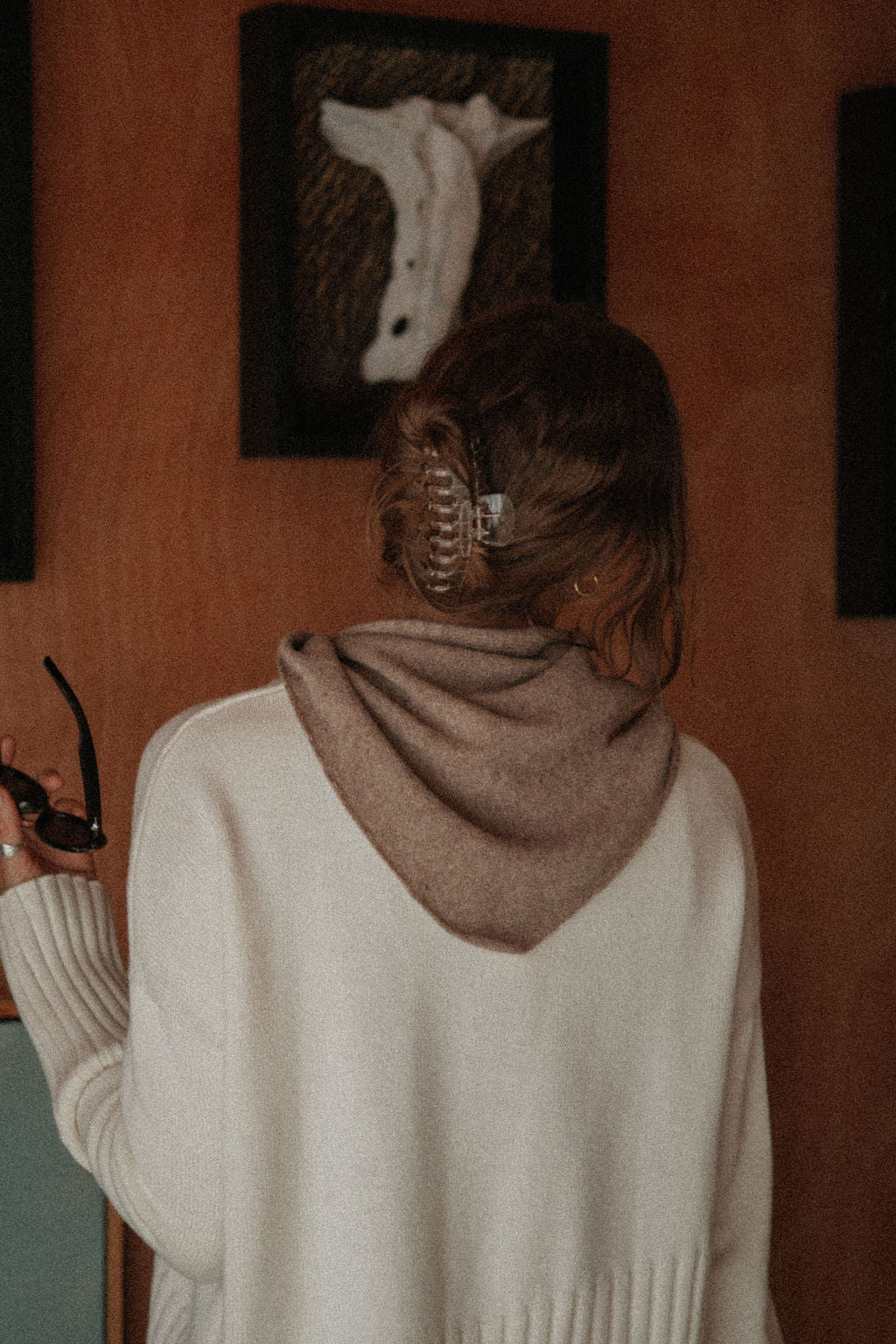 Person wearing a beige sweater with a brown hood in a room with a painting on the wall.