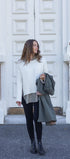Woman standing outside a building with large white doors, wearing a white sweater and black pants.
